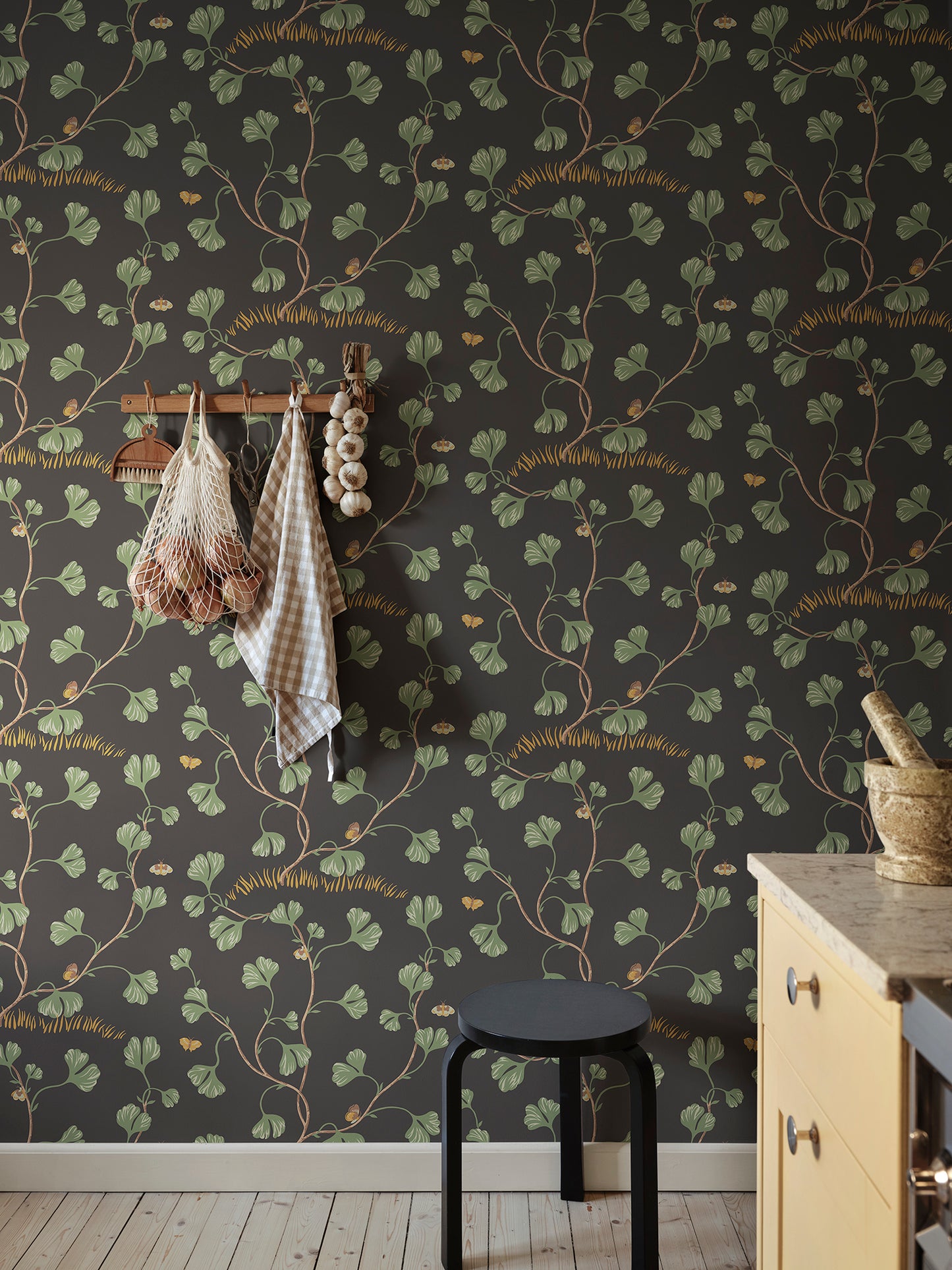 Ebba Charcoal is a sophisticated wallpaper featuring elegant ginkgo leaves and hidden butterflies on a striking dark background.