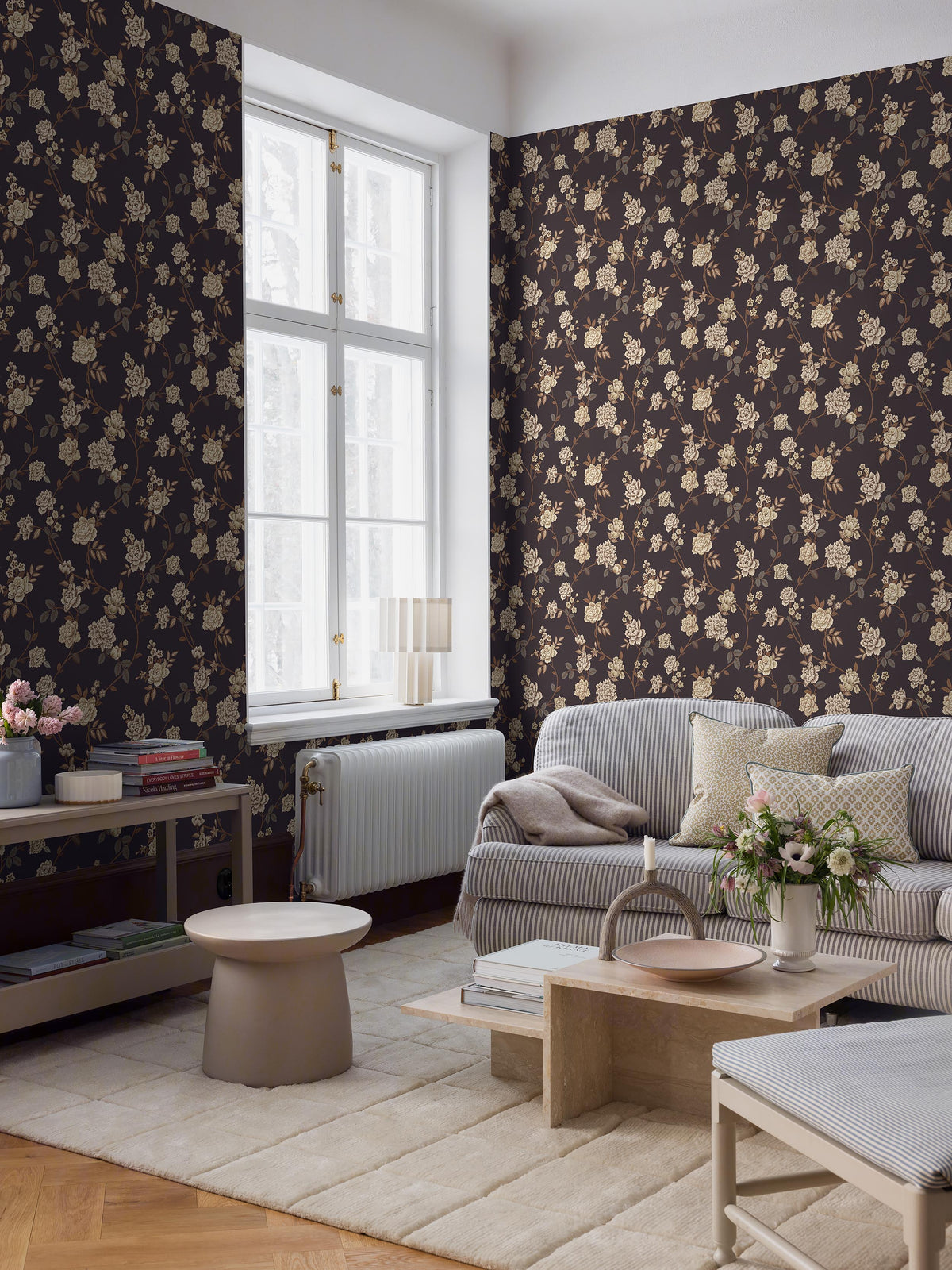 The dark, large-floral wallpaper Peony Garden creates a romantic yet slightly dramatic impression, with its delicate floral vines surface-printed in beige, grey and brown on a warm brown-black background.