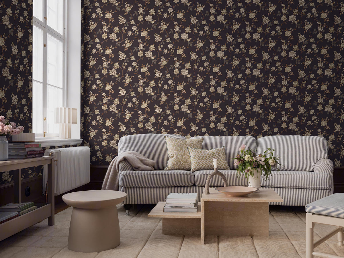 The dark, large-floral wallpaper Peony Garden creates a romantic yet slightly dramatic impression, with its delicate floral vines surface-printed in beige, grey and brown on a warm brown-black background.