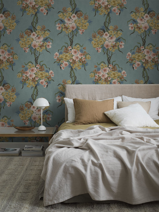 The modern floral wallpaper Colorful Flower showcases a dense pattern of stylized flowers and leaves in a muted yet intriguing color scheme: gray-green foliage with flowers in various shades of blue, rust pink, cream white, brown, and beige on a dark brown background.