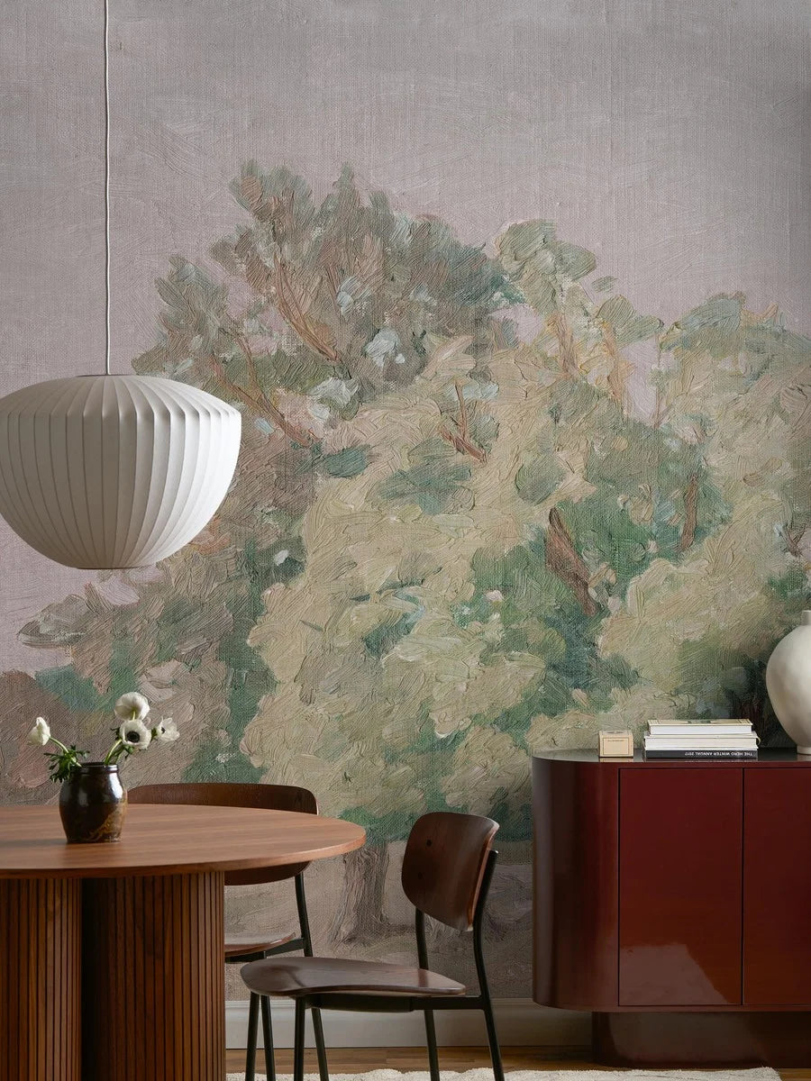 Like a large oil painting, the Hedgerow Landscape wallpaper stretches across the wall – an open landscape with softly shaped trees in hazy evening light.