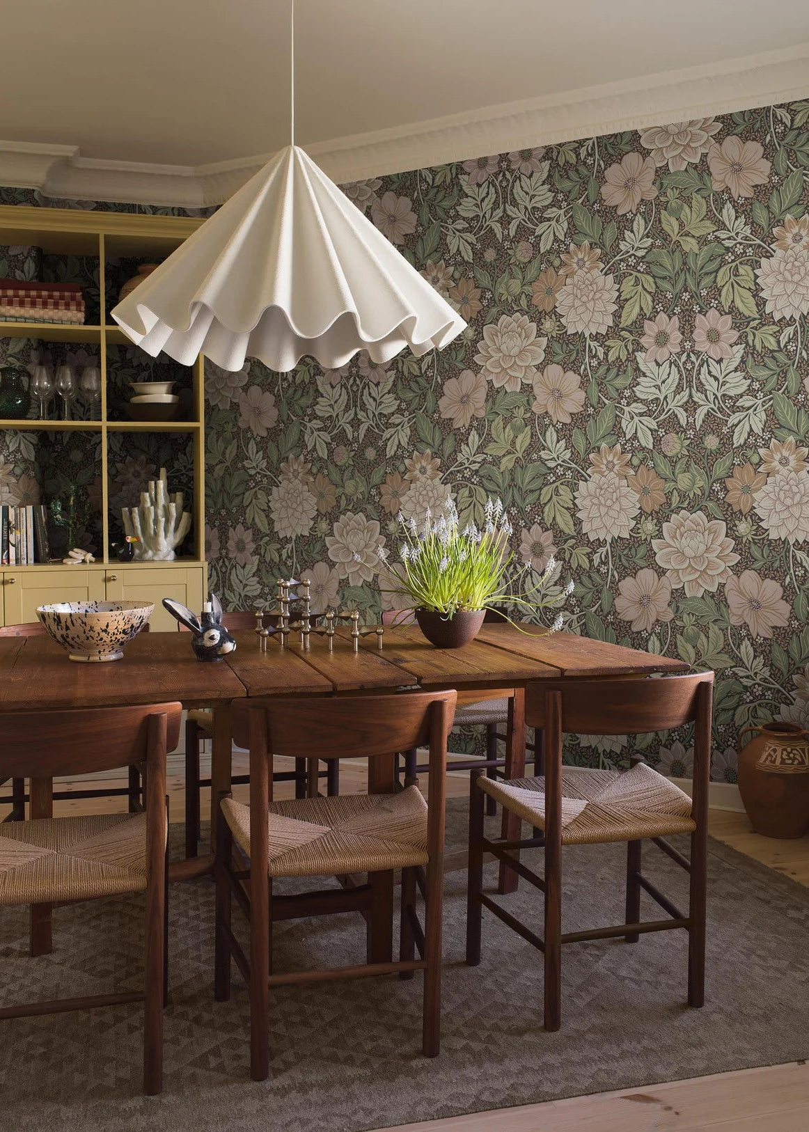 Dahlia Garden Grand mural Wallpaper's color palette is warm, soft, and inviting: pink, cream, light terracotta, and warm green shades on a dark background. The wallpaper is custom fit to your wall.