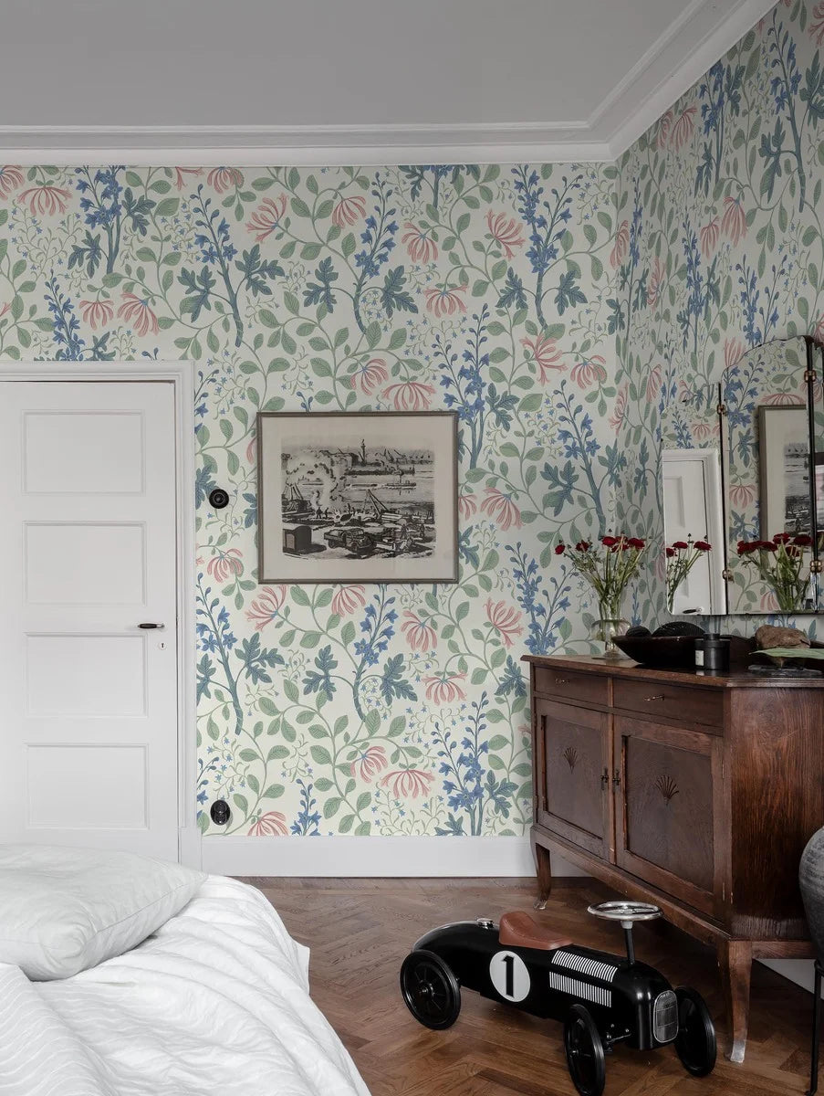 Across the light, large-floral Trädgårdsfröjd Grand wallpaper climbs a winding green foliage accented with blue delphiniums and pink honeysuckle on a cream-white background.