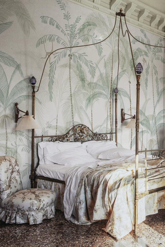 Hand-painted swaying palm trees with a vintage feel create a lush, airy scenography with monkeys swinging between the treetops. Wallpaper Louie, Light Green features a serene white background accented with delicate light green and soft beige hues.